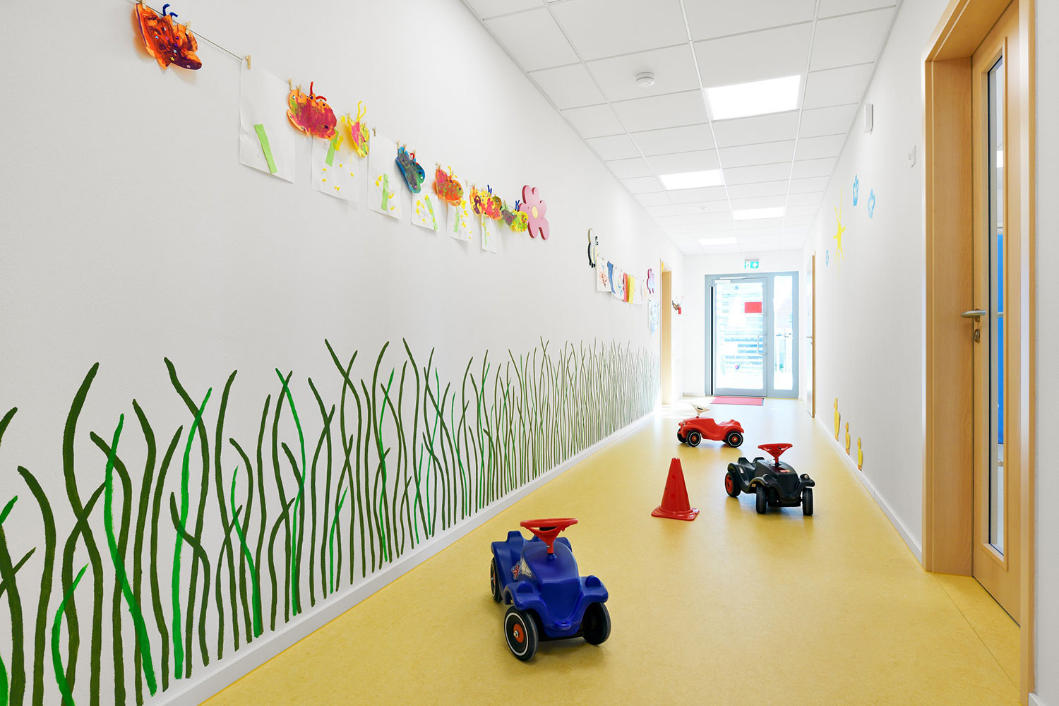 Flur einer Kindertagesstätte mit Bobycars, an der Wand sind Grashalme gemalt und es hängen selbstgemalte Bilder und Schmetterlinge an der Wand